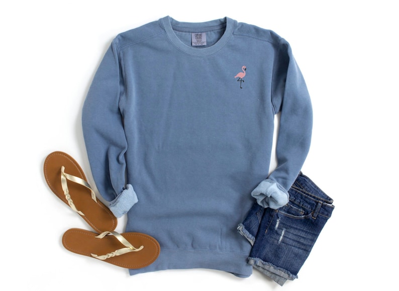 May include: A blue crew neck sweatshirt with a small pink flamingo embroidered on the chest. The sweatshirt is paired with denim shorts and brown sandals with gold straps, all on a white background.