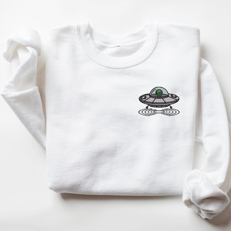 May include: White sweatshirt with a black and gray embroidered image of a UFO with a green alien inside. The UFO is hovering over a circle of ripples.