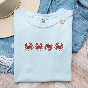 Könnte beinhalten: Hellblaues Comfort Colors T-Shirt mit vier gestickten roten Krebsen auf der Brust. Das Shirt ist gefaltet, mit einem sichtbaren Etikett. Ein Strohhut und Jeansshorts sind im Hintergrund.