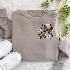 May include: A grey sweatshirt with a floral and butterfly design embroidered on the front. The design features three orange butterflies with black outlines and white flowers.