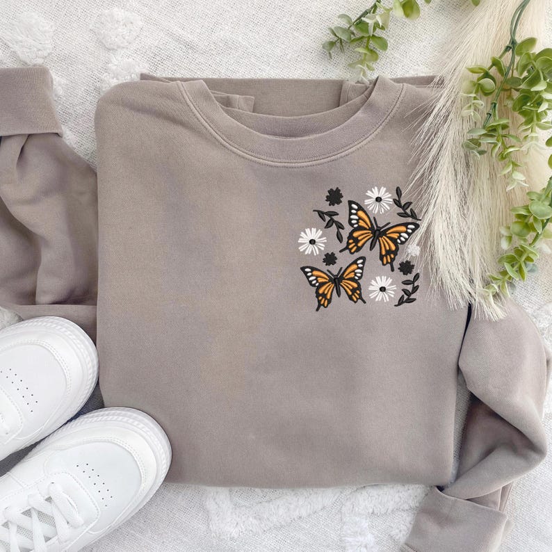 May include: A grey sweatshirt with a floral and butterfly design embroidered on the front. The design features three orange butterflies with black outlines and white flowers.