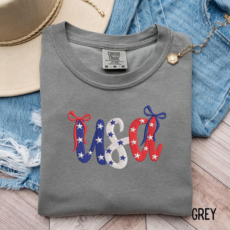 Pu&ograve; includere: Una felpa grigia girocollo con la scritta "USA" ricamata in rosso, bianco e blu, decorata con stelle e fiocchi. La maglietta &egrave; realizzata in un materiale morbido ed &egrave; etichettata "Comfort Colors". La parola "GREY" &egrave; stampata in basso a destra.