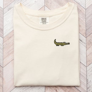 Puede incluir: Una camiseta beige con un pequeño cocodrilo bordado en la parte delantera. La camiseta tiene cuello redondo y está hecha de una tela suave y cómoda. La etiqueta de la marca dice "COMFORT COLORS" con el texto "HEAVYWEIGHT FLEECE" y "100% COTTON" debajo.
