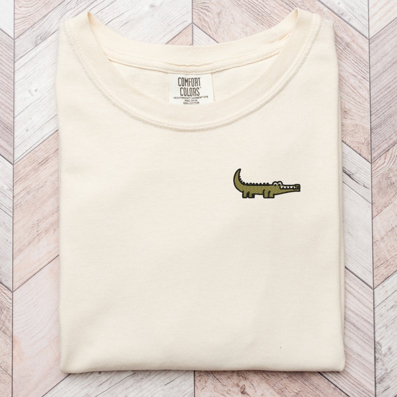 Peut inclure: Un t-shirt beige avec un petit alligator brod&eacute; sur le devant. Le t-shirt a un col rond et est fait d'un tissu doux et confortable. L'&eacute;tiquette de la marque indique "COMFORT COLORS" avec le texte "HEAVYWEIGHT FLEECE" et "100% COTTON" en dessous.