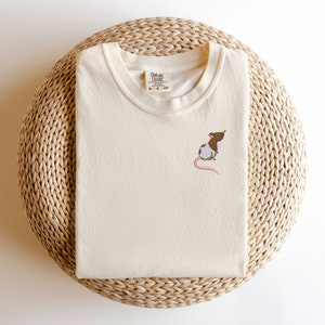 May include: A cream colored t-shirt with a brown and white embroidered rat on the front. The t-shirt has a crew neck and the text "Common Colors" on the tag.