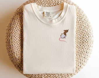 May include: A cream colored t-shirt with a brown and white embroidered rat on the front. The t-shirt has a crew neck and the text "Common Colors" on the tag.