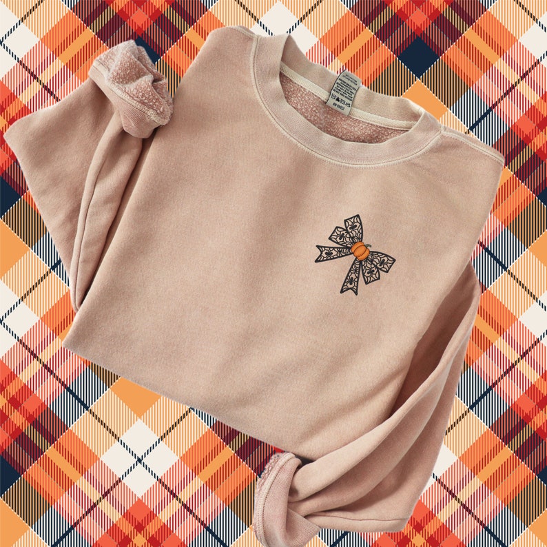 Puede incluir: Una sudadera rosa claro con un lazo negro bordado con una calabaza en el centro. La sudadera est&aacute; sobre un fondo a cuadros.