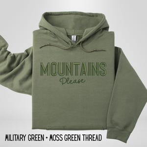 May include: An olive green hoodie with the text "MOUNTAINS Please" in green. The hoodie has a drawstring and a front pocket. The bottom of the image reads "MILITARY GREEN + MOSS GREEN THREAD".