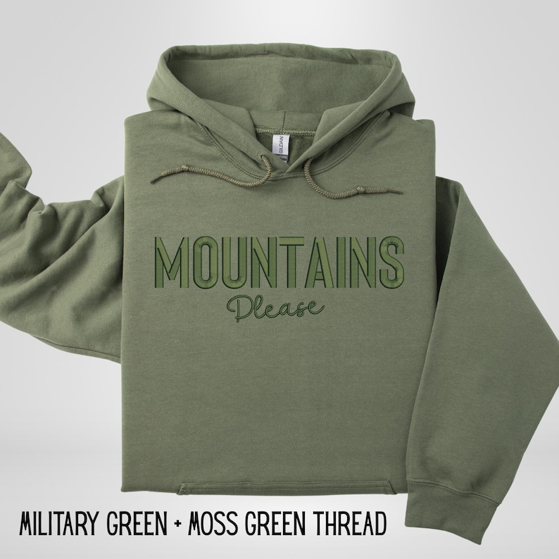 May include: An olive green hoodie with the text "MOUNTAINS Please" in green. The hoodie has a drawstring and a front pocket. The bottom of the image reads "MILITARY GREEN + MOSS GREEN THREAD".