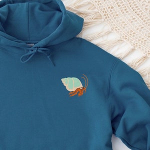 Puede incluir: Una sudadera con capucha azul turquesa con una pequeña imagen bordada de un cangrejo ermitaño en una concha azul claro.