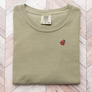 Puede incluir: Una camiseta verde doblada con una peque&ntilde;a mariquita roja bordada en la parte delantera. La camiseta tiene una etiqueta que dice "Comfort Colors" y "S - P - CH".