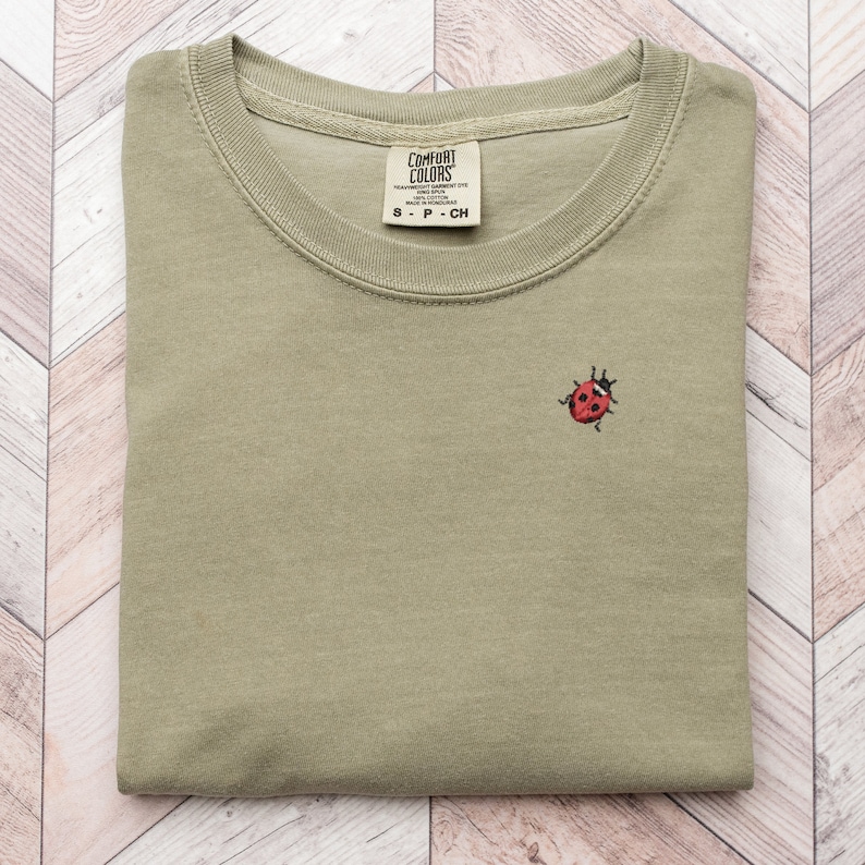 Puede incluir: Una camiseta verde doblada con una peque&ntilde;a mariquita roja bordada en la parte delantera. La camiseta tiene una etiqueta que dice "Comfort Colors" y "S - P - CH".