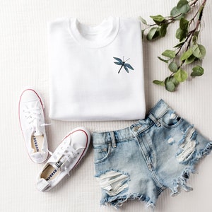 May include: A white sweatshirt with a blue dragonfly embroidered on the front, a pair of white sneakers, and a pair of light blue denim shorts with distressed details.