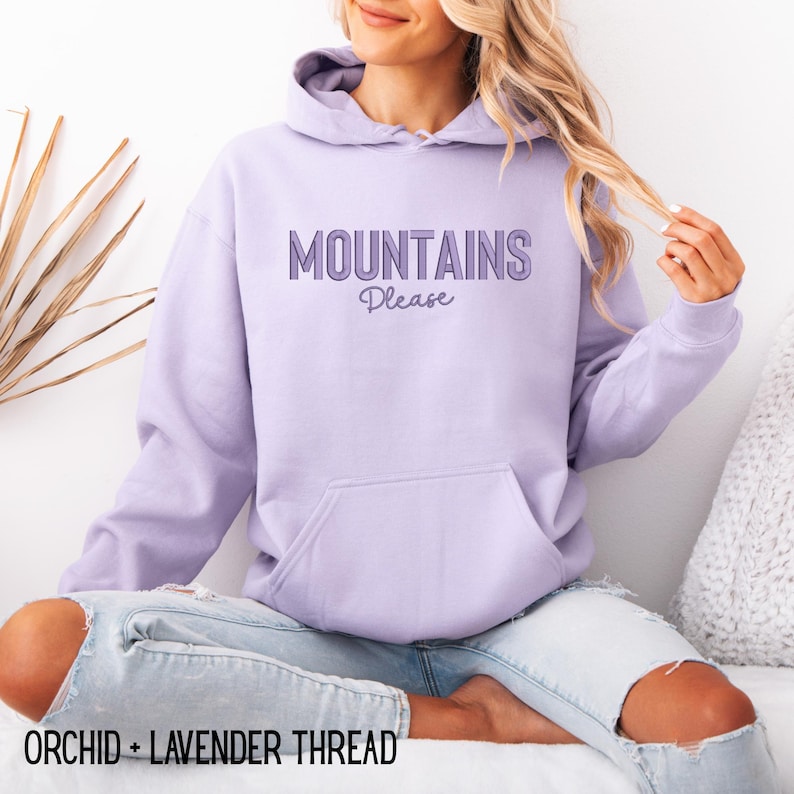 May include: A light purple hoodie with the text "MOUNTAINS Please" in a darker purple font. The hoodie has a front pocket and a hood. The words "ORCHID + LAVENDER THREAD" are at the bottom of the image.