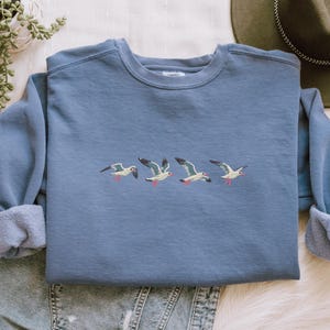Puede incluir: Una sudadera azul polvorienta con un dise&ntilde;o bordado de cuatro gaviotas en vuelo. Las aves tienen cuerpos blancos, alas grises y detalles rojos en sus picos y patas. La sudadera tiene cuello redondo y mangas largas.