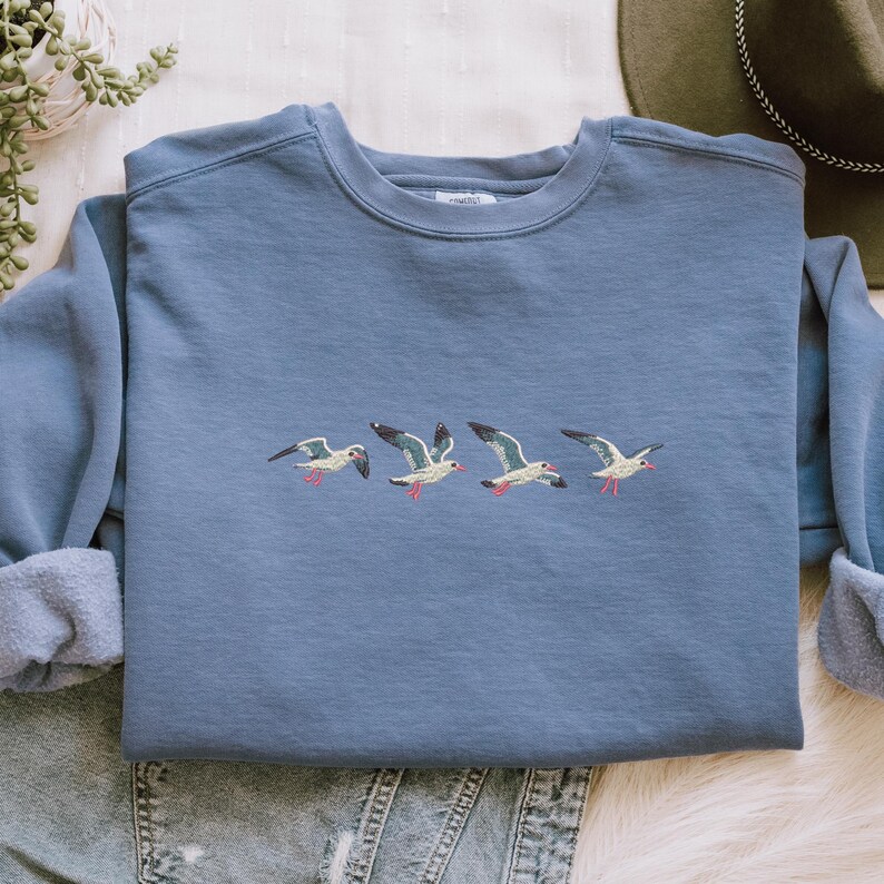 Puede incluir: Una sudadera azul polvorienta con un dise&ntilde;o bordado de cuatro gaviotas en vuelo. Las aves tienen cuerpos blancos, alas grises y detalles rojos en sus picos y patas. La sudadera tiene cuello redondo y mangas largas.
