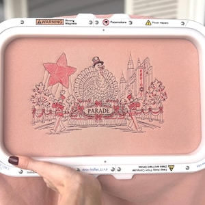 Peut inclure: Un cadre à broder rectangulaire blanc contenant un tissu rose avec une scène brodée à la machine représentant une dinde en haut-de-forme sur un char de parade, entourée de deux musiciens, une grande étoile et des bâtiments avec le texte RADIO CITY.