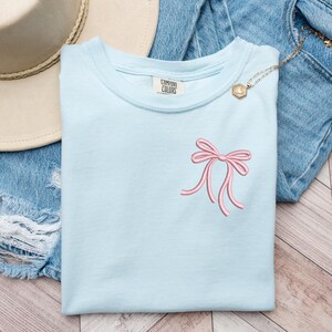 以下が含まれることがあります： 薄い青色のTシャツに、ピンクのリボンが刺繍されています。Tシャツには、「COMFORT COLORS」と書かれた白いタグが付いています。