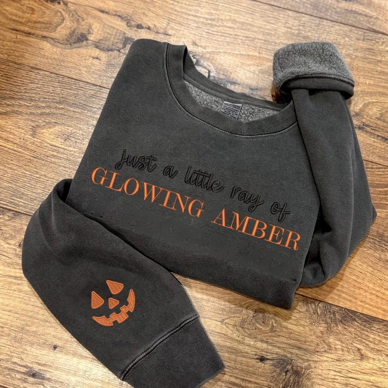 May include: Dark gray sweatshirt with the text "Just a little ray of GLOWING AMBER" in orange script. The sleeve features an embroidered orange jack-o'-lantern. The sweatshirt has a crew neck and a fleece lining.