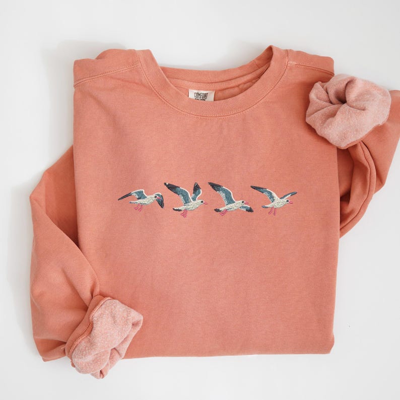 Puede incluir: Una sudadera de color coral con gaviotas bordadas en vuelo. La parte superior de manga larga tiene cuello redondo y una scrunchie a juego. Las gaviotas est&aacute;n representadas en tonos de azul, blanco y gris.