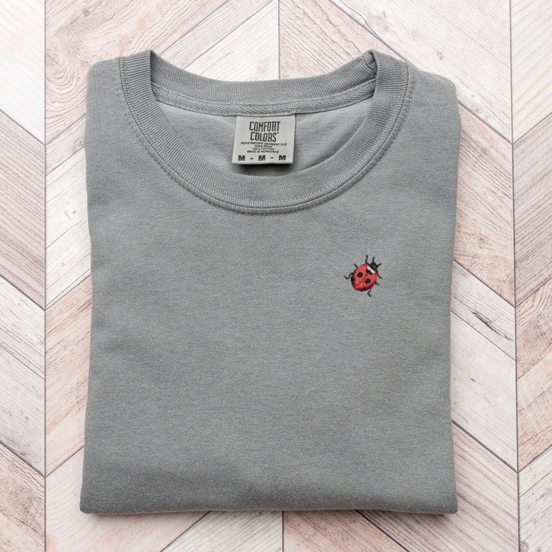 Puede incluir: Una camiseta gris con una peque&ntilde;a mariquita roja bordada en la parte delantera. La camiseta tiene una etiqueta de Comfort Colors con la talla M.