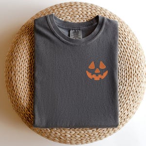 May include: Dark grey t-shirt featuring an orange jack-o'-lantern face embroidered on the chest. The shirt is folded on a round, woven wicker surface. The t-shirt has a crew neck and short sleeves, ideal for Halloween.