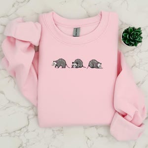 May include: A pink sweatshirt with a graphic of three opossums embroidered on the front.