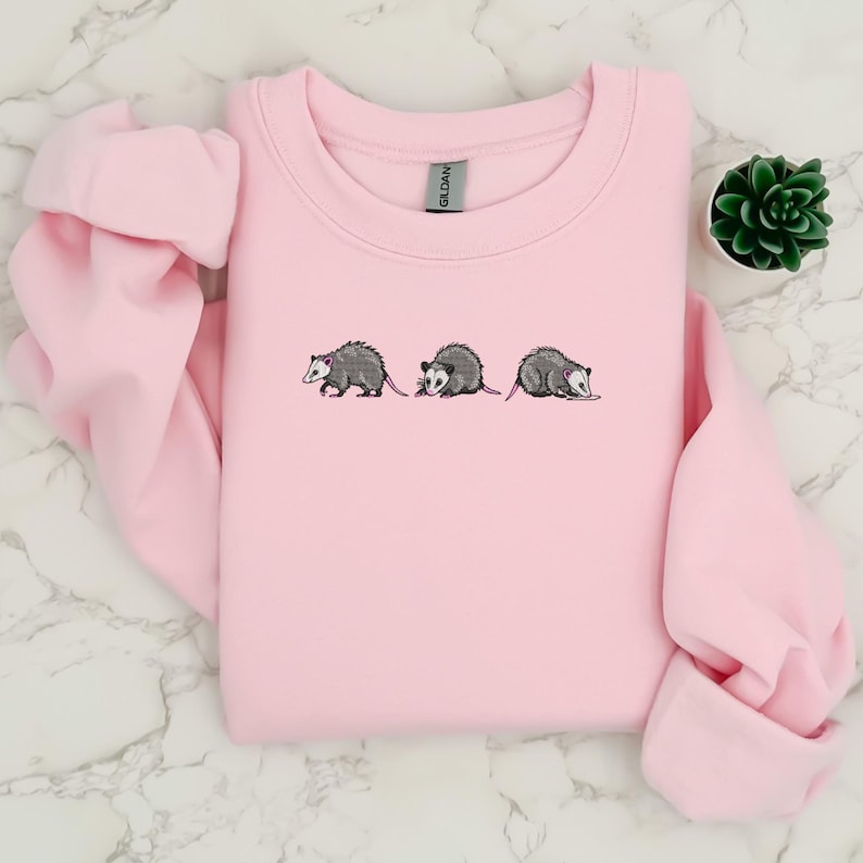 May include: A pink sweatshirt with a graphic of three opossums embroidered on the front.