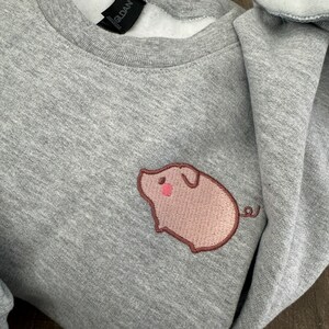 K&ouml;nnte beinhalten: Grauer Pullover mit einem gestickten rosa Schwein auf der linken Brustseite. Das Schwein hat eine rosa Nase und einen braunen Schwanz.