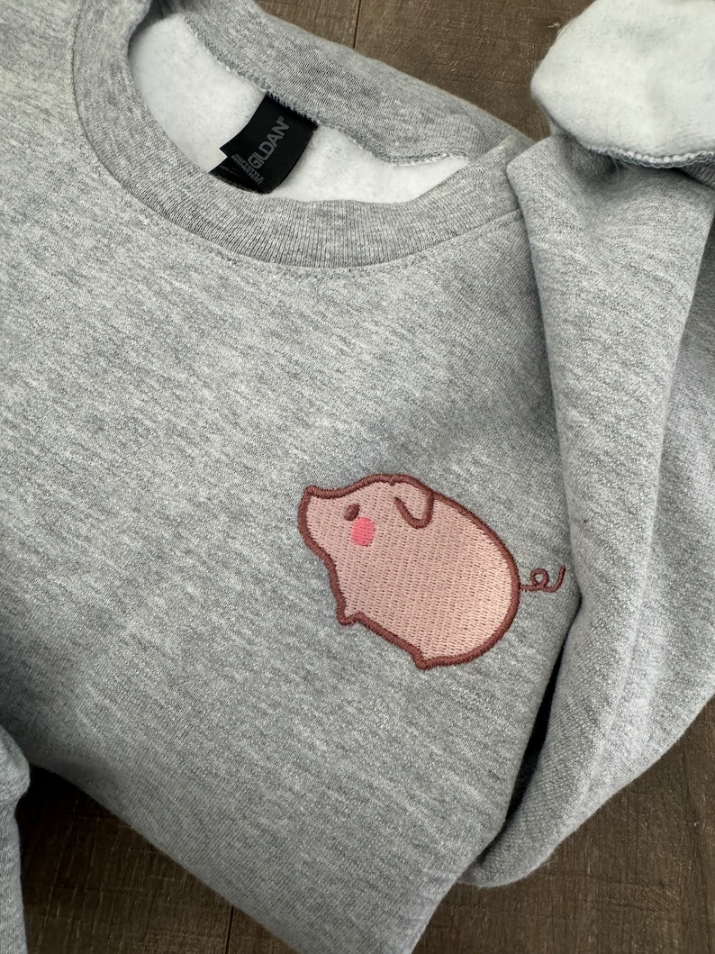 K&ouml;nnte beinhalten: Grauer Pullover mit einem gestickten rosa Schwein auf der linken Brustseite. Das Schwein hat eine rosa Nase und einen braunen Schwanz.