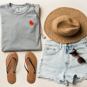 Puede incluir: Una camiseta gris con un gr&aacute;fico de sand&iacute;a, un par de chanclas marrones, un sombrero de paja y pantalones cortos de mezclilla azul claro con un par de gafas de sol en el bolsillo.