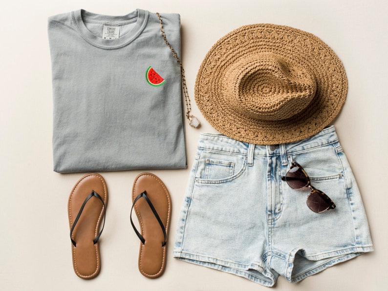 Puede incluir: Una camiseta gris con un gr&aacute;fico de sand&iacute;a, un par de chanclas marrones, un sombrero de paja y pantalones cortos de mezclilla azul claro con un par de gafas de sol en el bolsillo.