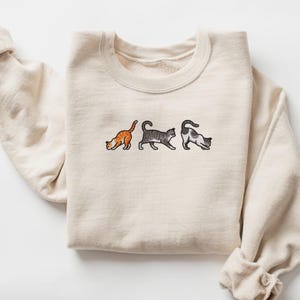 May include: A cream-colored sweatshirt with three embroidered cats in a row. The cats are orange, gray, and black and white.