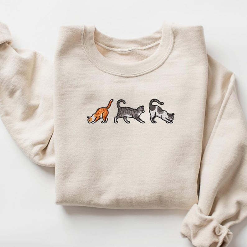 May include: A cream-colored sweatshirt with three embroidered cats in a row. The cats are orange, gray, and black and white.
