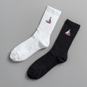 May include: Two pairs of crew socks, one white and one black. Each sock features an embroidered sailboat design in red, white, and blue. The socks are ribbed and made of a soft material, perfect for casual wear.