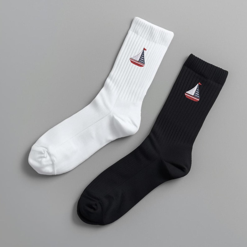 May include: Two pairs of crew socks, one white and one black. Each sock features an embroidered sailboat design in red, white, and blue. The socks are ribbed and made of a soft material, perfect for casual wear.