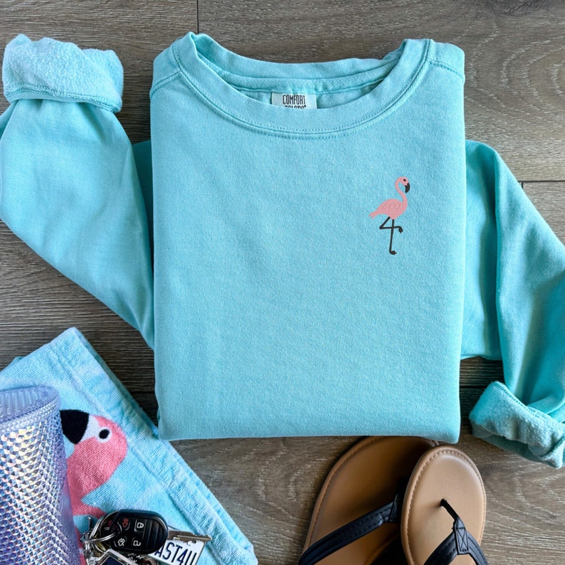 May include: A light blue sweatshirt with a small pink flamingo embroidered on the chest. The long-sleeved shirt is folded neatly, with the sleeves rolled up. A matching towel, flip-flops, and keys are also visible.