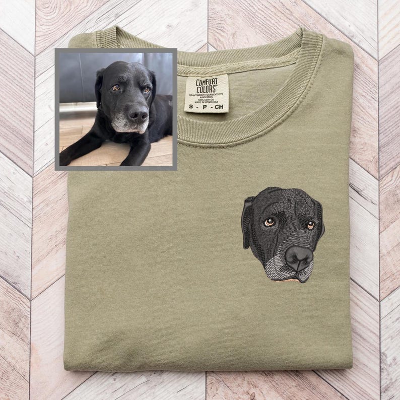 May include: A sage green Comfort Colors t-shirt featuring an embroidered black dog head on the front. A small framed photo of a black dog is in the upper left corner. The shirt has a crew neck and short sleeves.