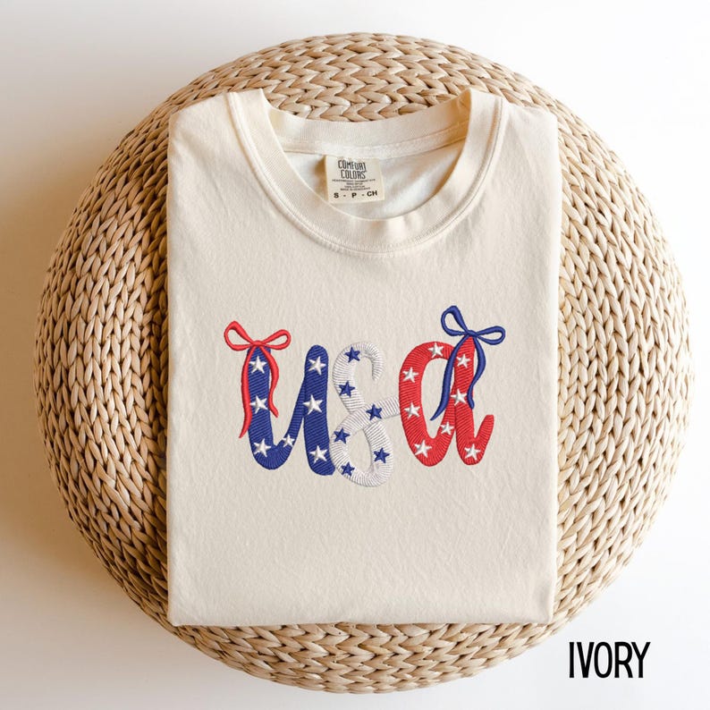 Pu&ograve; includere: T-shirt color avorio con la parola "USA" ricamata in rosso, bianco e blu, ornata di stelle e fiocchi. La maglietta &egrave; realizzata in un materiale morbido ed &egrave; esposta su una superficie tessuta. L'etichetta recita "Comfort Colors".
