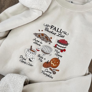 May include: Cream-colored sweatshirt with an embroidered "FALL Bucket List" design. The design includes illustrations of leaves, apples, a pie, a pumpkin, and a cup of apple cider. The sweatshirt has a soft, textured collar.
