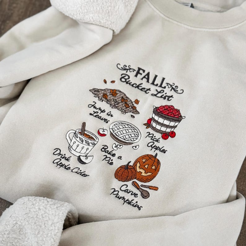 May include: Cream-colored sweatshirt with an embroidered "FALL Bucket List" design. The design includes illustrations of leaves, apples, a pie, a pumpkin, and a cup of apple cider. The sweatshirt has a soft, textured collar.