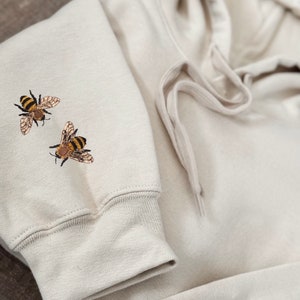 May include: A light beige hoodie with two embroidered bees on the left sleeve.