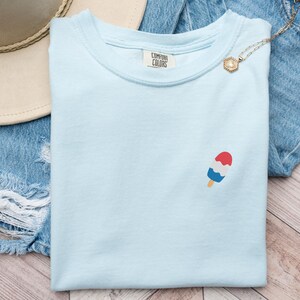 May include: Light blue Comfort Colors t-shirt with a small popsicle graphic in red, white, and blue. The shirt is displayed on denim shorts and a wooden surface. A gold necklace and a straw hat are also in the image.