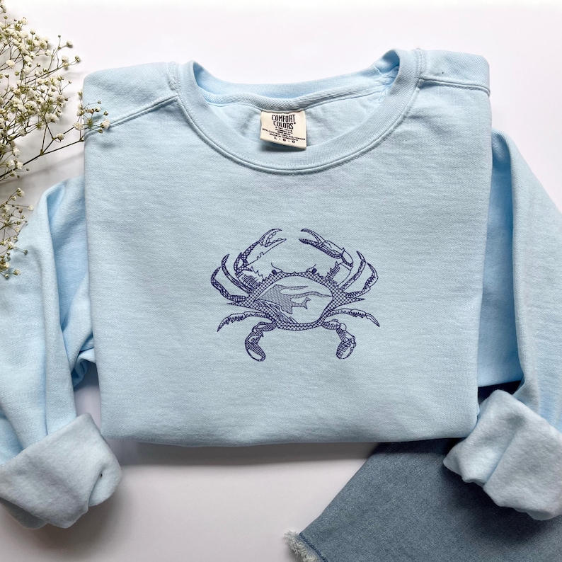 K&ouml;nnte beinhalten: Hellblaues Sweatshirt mit einem marineblauen Krabbenmuster, auf die Vorderseite gestickt. Das Sweatshirt hat einen Rundhalsausschnitt und lange &Auml;rmel. Das Comfort Colors-Etikett ist am Ausschnitt sichtbar. Das Sweatshirt liegt auf einer blauen Jeans.