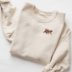 May include: A cream coloured sweatshirt with a small embroidered tiger on the left chest.