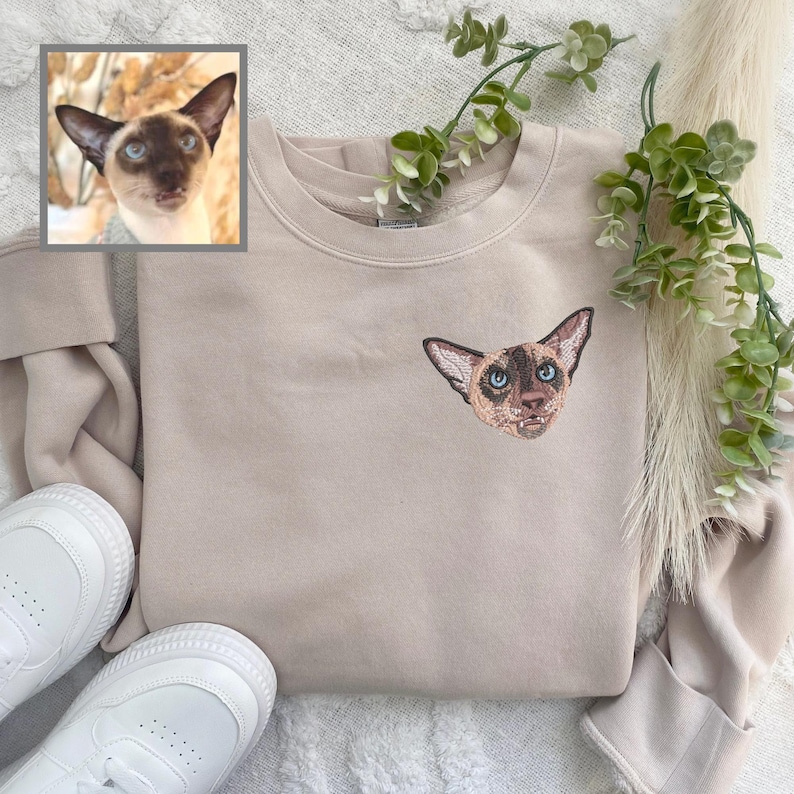 Puede incluir: Una sudadera beige con una imagen bordada personalizada de la cara de un gato siam&eacute;s. El gato tiene ojos azules y un pelaje marr&oacute;n claro y blanco.