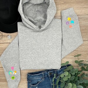 Peut inclure: Un sweat &agrave; capuche gris avec des patchs color&eacute;s sur les manches. Les patchs sont en forme de c&oelig;ur, d'arc-en-ciel, de tr&egrave;fle &agrave; quatre feuilles et d'&eacute;toile.