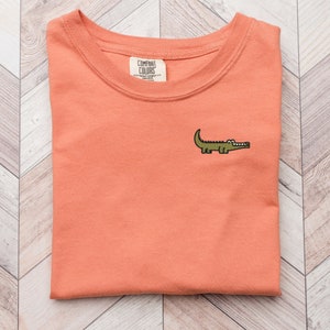 Peut inclure: Un t-shirt de couleur corail avec un petit alligator brod&eacute; sur le devant. Le t-shirt a un col rond et est fait d'un tissu doux et confortable. L'&eacute;tiquette de la marque indique "COMFORT COLORS" avec le texte "HEAVYWEIGHT GARMENT DYE" et "MADE IN JAPAN".