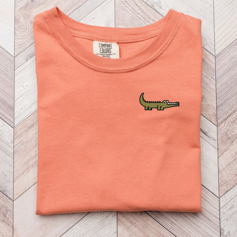 Peut inclure: Un t-shirt de couleur corail avec un petit alligator brod&eacute; sur le devant. Le t-shirt a un col rond et est fait d'un tissu doux et confortable. L'&eacute;tiquette de la marque indique "COMFORT COLORS" avec le texte "HEAVYWEIGHT GARMENT DYE" et "MADE IN JAPAN".