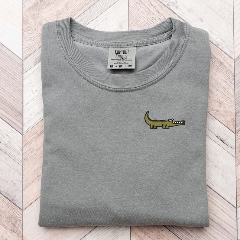 Peut inclure: Un t-shirt gris avec un petit alligator brod&eacute; sur le devant. Le t-shirt a un col rond et est pli&eacute; en deux. L'&eacute;tiquette sur le t-shirt indique "Comfort Colors".